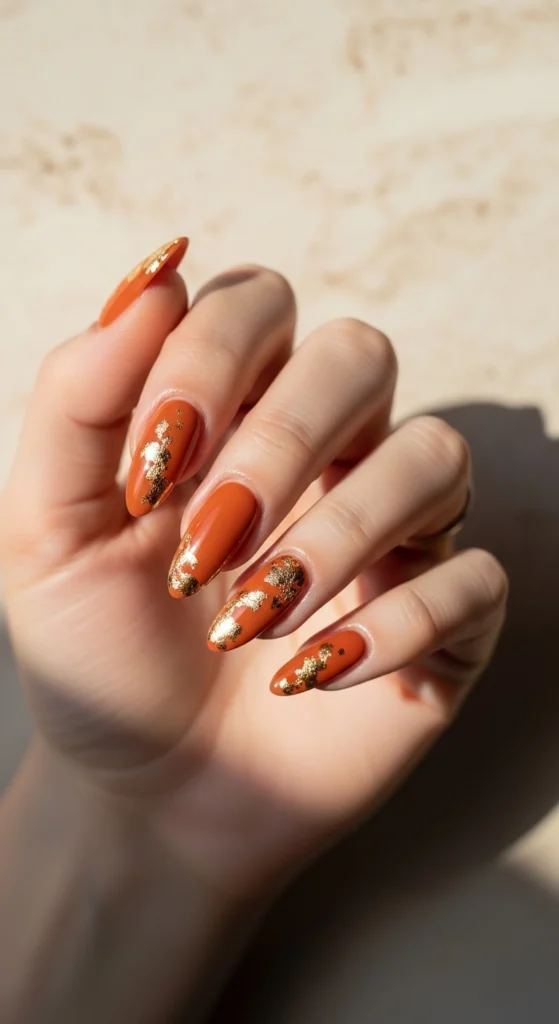 15 Fall Nail Art Ideas for 2026: Warm Tones, Foils & Seasonal Styles 5 Almond-shaped nails painted in burnt orange with delicate gold accents, on a soft beige marble background.