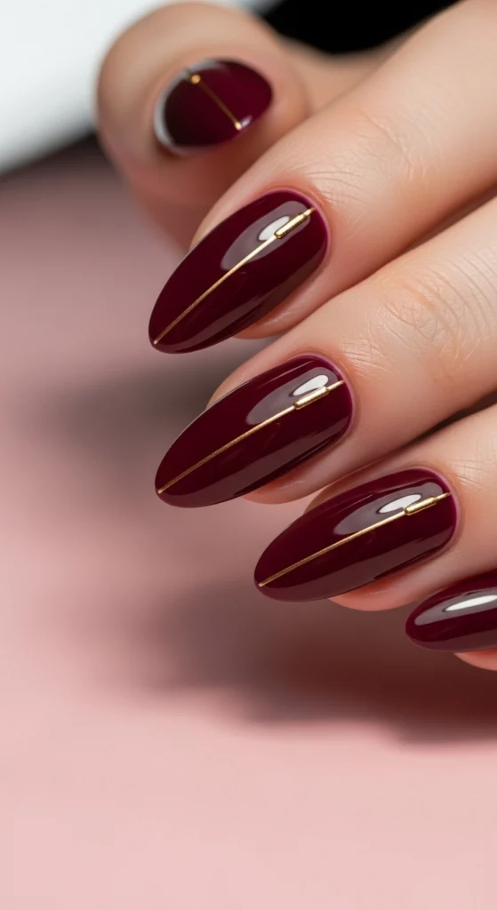 15 Fall Nail Art Ideas for 2026: Warm Tones, Foils & Seasonal Styles 6 Almond-shaped nails in deep wine glossy polish with thin metallic gold lines, photographed against a blush pink background under soft beauty lighting.
