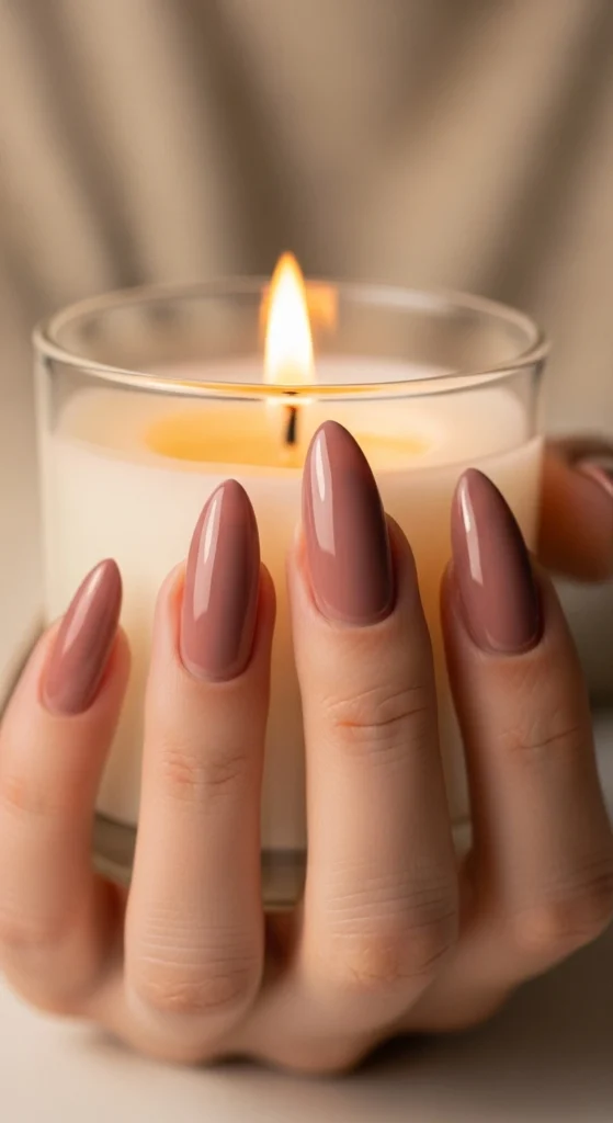 Glossy mauve almond nails holding a small scented candle on a beige background.