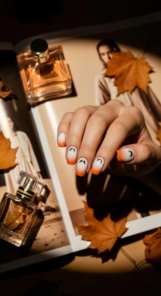 Flat lay of short rounded iridescent gradient nails white to orange with chrome moon accents, styled with perfume and fall leaves.