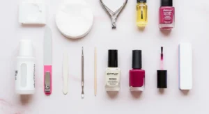 Manicure at Home. A bright, clean, and inviting flat lay photograph showcasing all the essential tools for an at-home manicure. Arrange neatly on a soft, light-colored (e.g., pastel pink or white marble) background: a bottle of nail polish remover, cotton pads, a nail clipper, a nail file, a cuticle pusher, a small bottle of cuticle oil, a base coat, a vibrant bottle of nail polish (e.g., a trending shade from Fall 2025 like deep burgundy), a glossy top coat, and a nail buffer. Some tools can be slightly out of focus in the background to add depth.