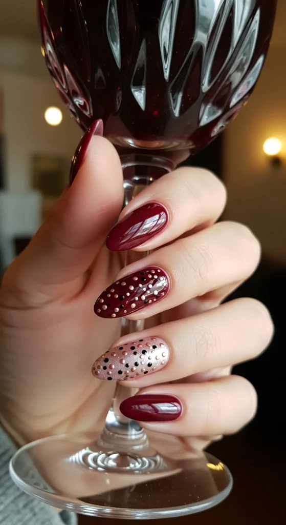 Glossy burgundy almond nails with scattered rose-gold and black dots, wine glass in the background.