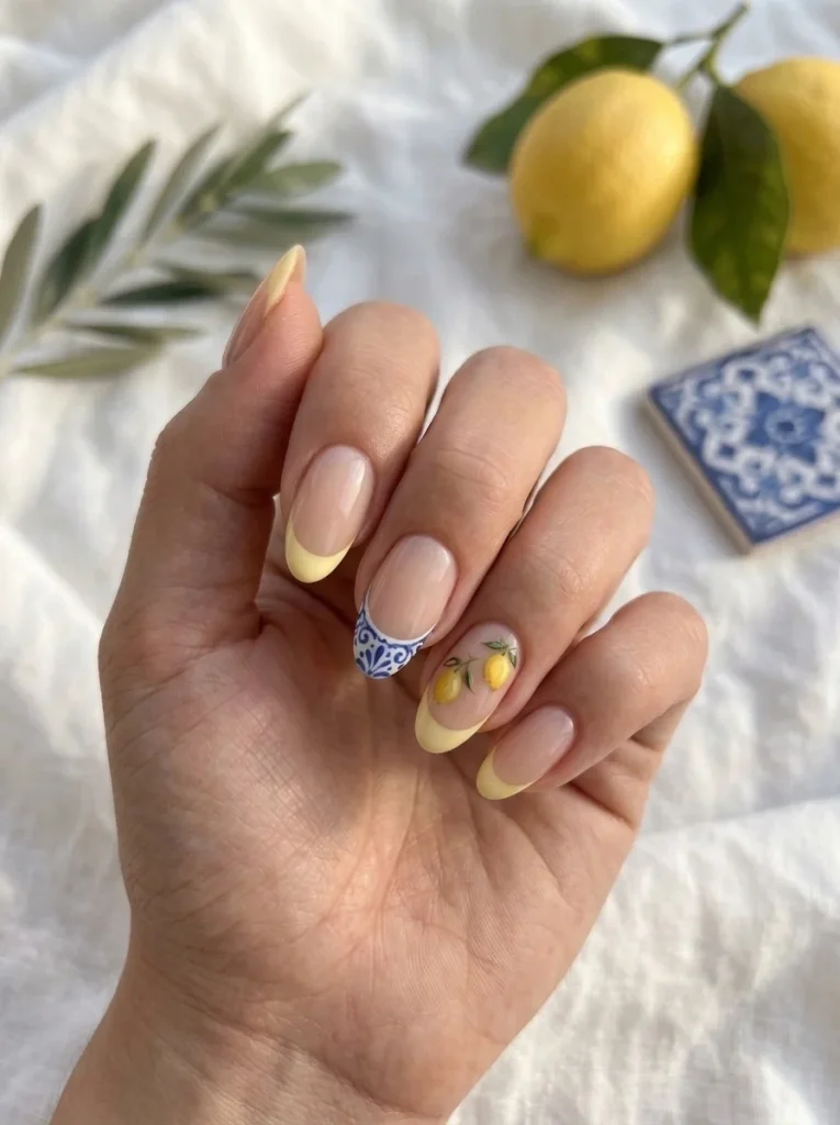 Amalfi Lemon Tips manicure with buttery yellow French tips, delicate hand-painted lemons, and a subtle Mediterranean ceramic accent on almond nails.