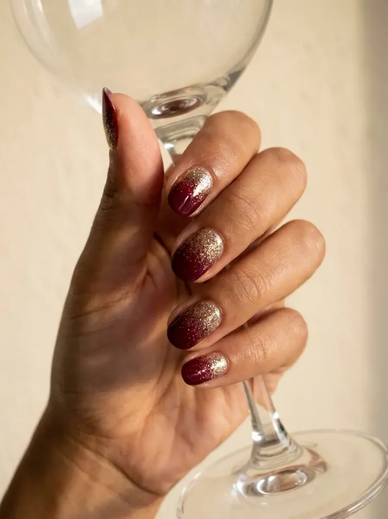 Short oval nails with gold glitter at the base fading into glossy burgundy tips on an olive skin tone