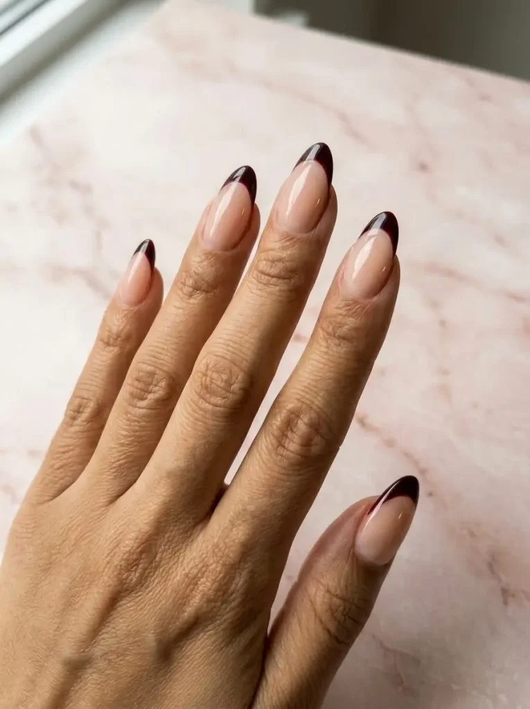 Almond nails with a sheer nude base and slim black cherry French tips, on a medium skin tone