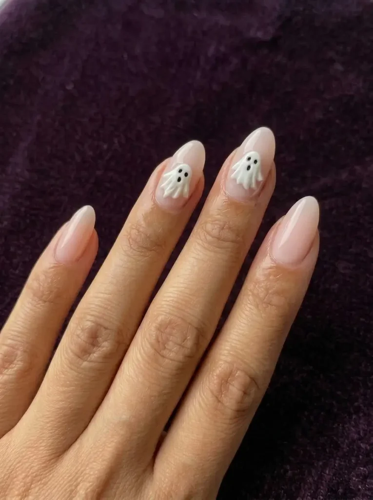 Milky pink almond nails with small white ghosts on the ring and middle nails, on a medium skin tone against dark plum velvet