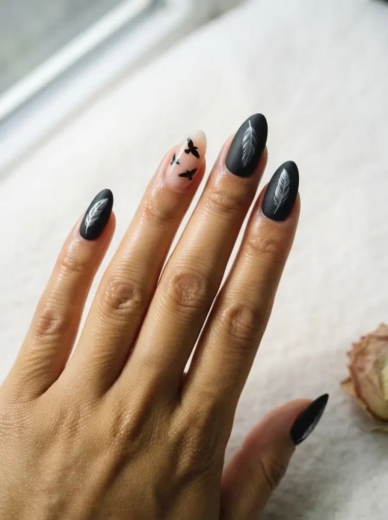 Matte black almond nails with silver feather art and one nude nail with black ravens in flight, on a medium skin tone