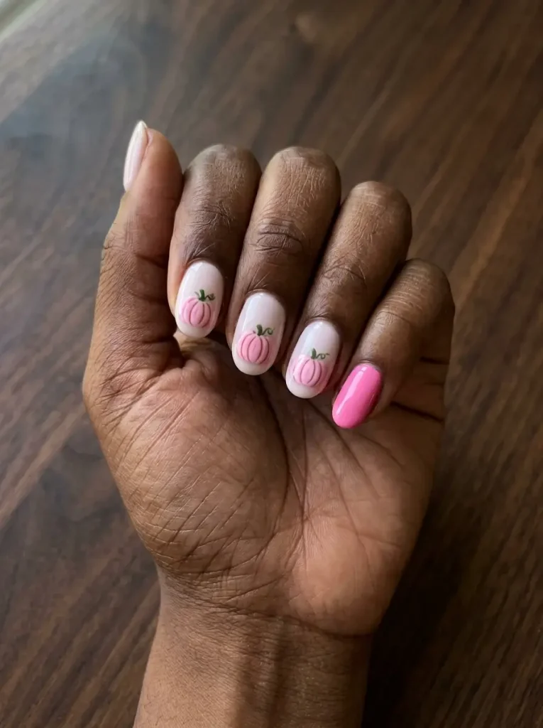 Short oval nails with pastel pink pumpkin art on a milky base and one solid bubblegum pink nail, on a deep skin tone