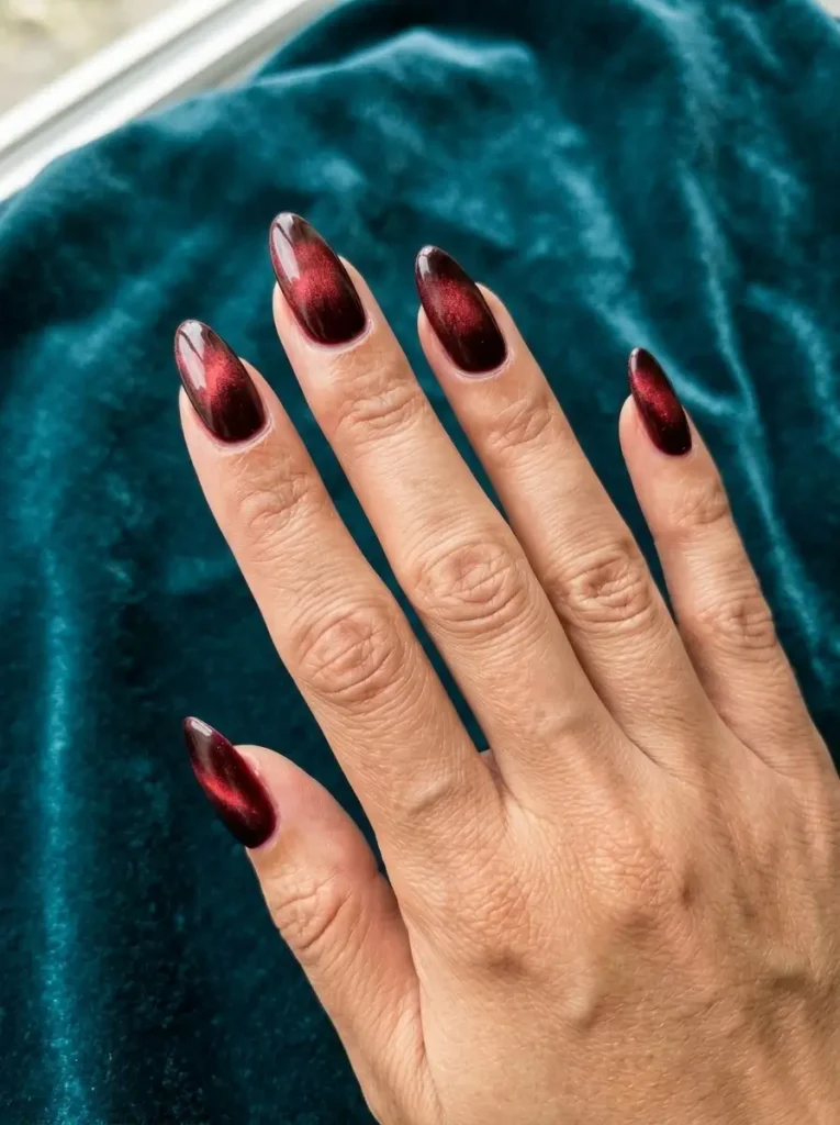 Vampire red velvet cat-eye almond nails on a hand against deep teal velvet fabric.