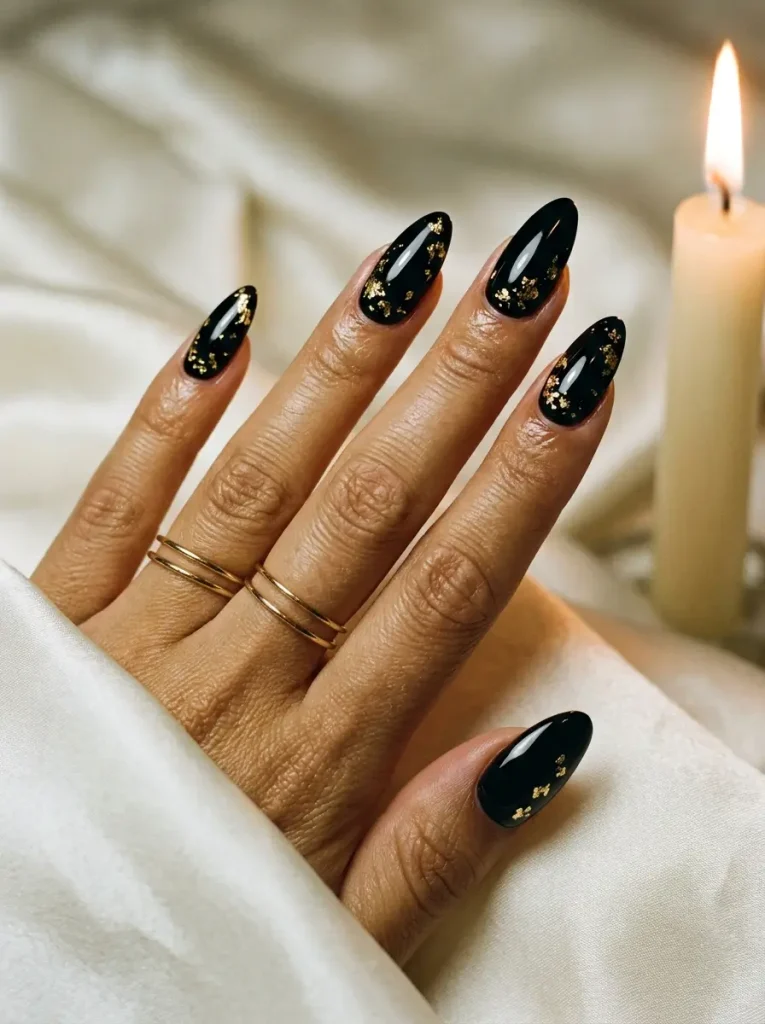Long almond nails in glossy black with flakes of gold foil near the cuticle, on a medium skin tone