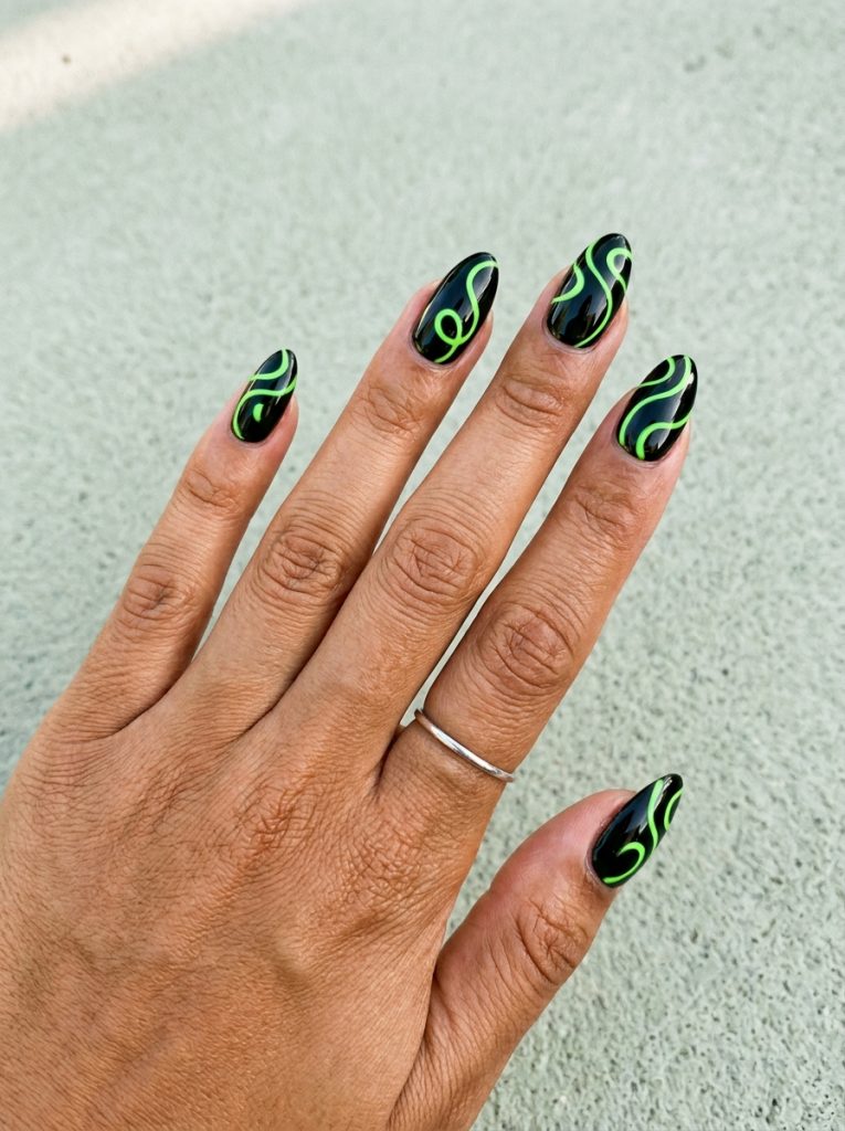 Medium almond nails in glossy black with thin neon green swirl lines, on a tan skin tone