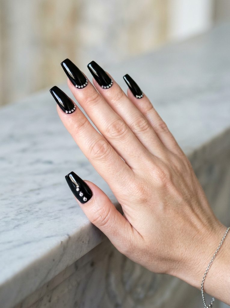 Long coffin nails in glossy black with a row of small silver studs along the cuticle, on a fair skin tone