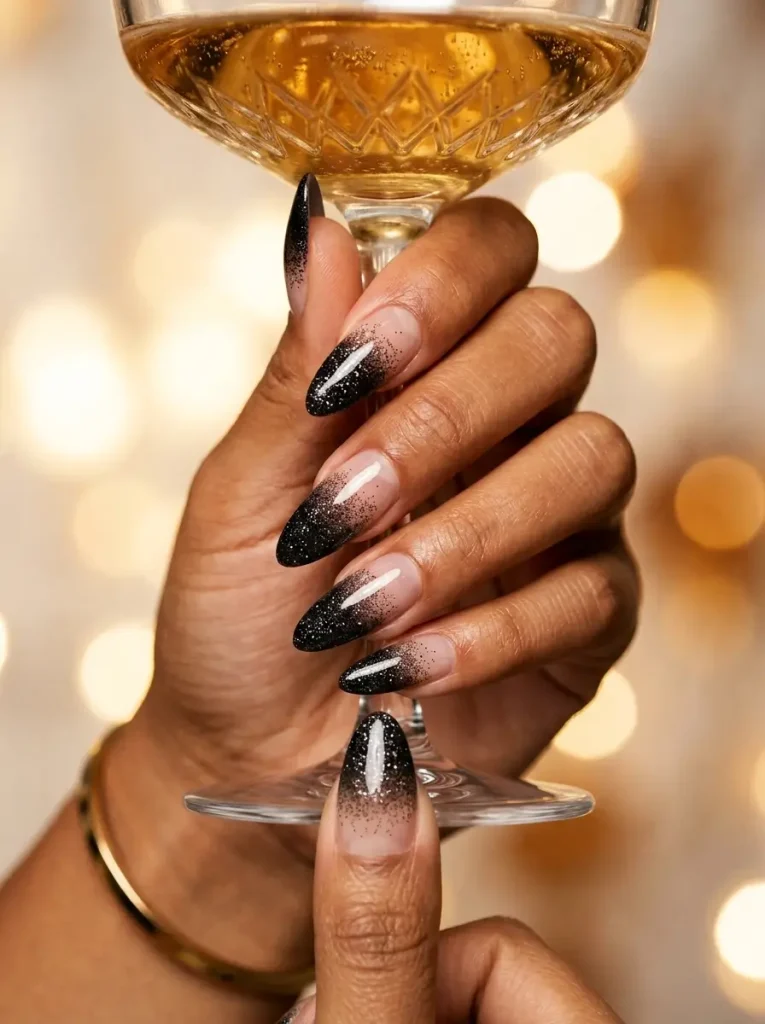 Long almond nails with fine black glitter dense at the tip and fading to sheer at the cuticle, on a tan skin tone