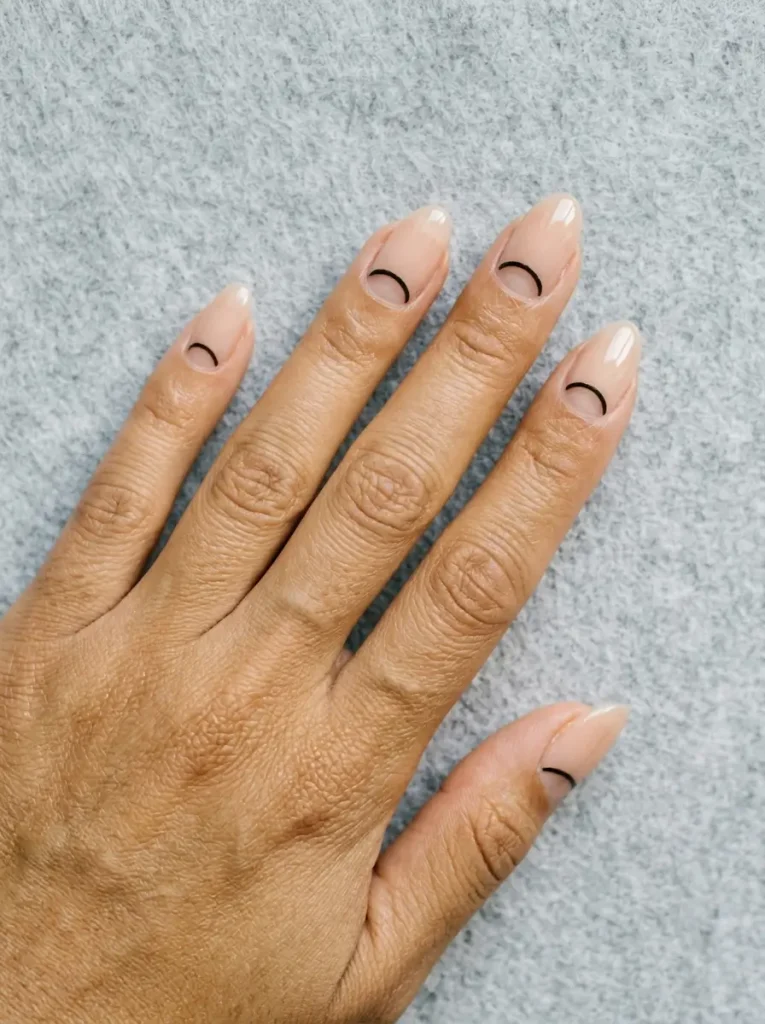 Oval nails with a sheer nude base and a bare half-moon at the cuticle outlined in a thin black crescent, on a medium skin tone
