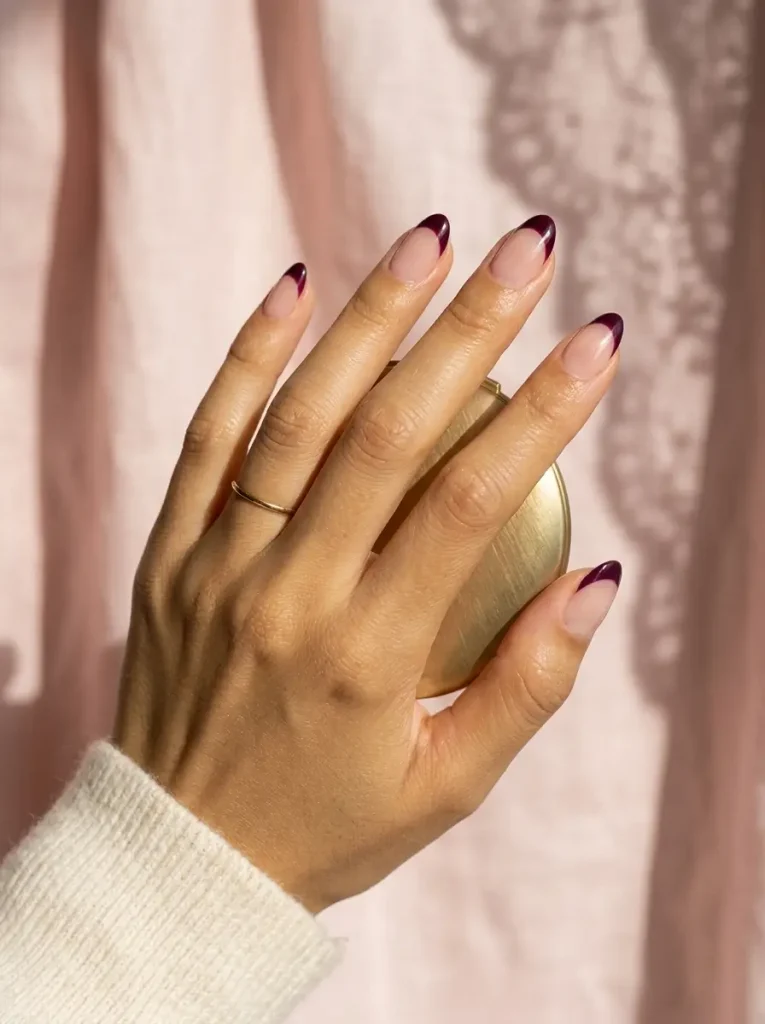Short almond nails with a sheer nude base and deep burgundy French tips, on a medium skin tone