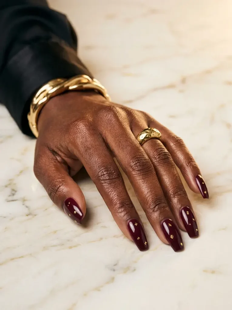 Medium coffin nails in glossy burgundy with scattered gold foil flakes, on a deep skin tone