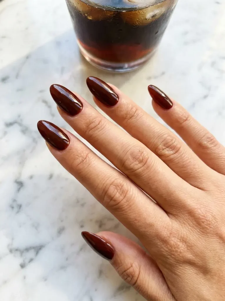 Long almond nails in glossy cherry cola brown, a deep red-brown shade, on a light skin tone