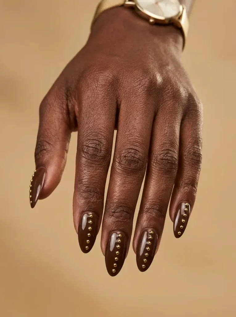 28 Fall Nail Designs for 2026, From Simple to Classy 4 Long almond glossy chocolate nails with a single vertical line of tiny gold studs on every nail, on a deep skin tone