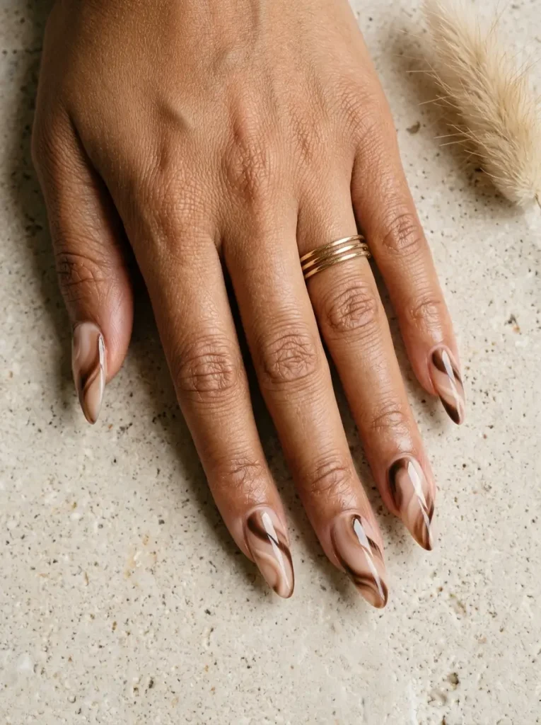 Long almond nails with swirls of espresso, mocha and cream across a nude base, on a tan skin tone