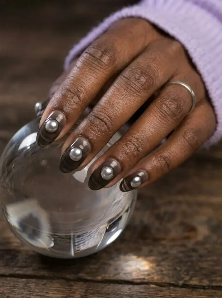 Oval nails with a smoky sheer black jelly base and a round iridescent chrome orb in the center, on a deep skin tone