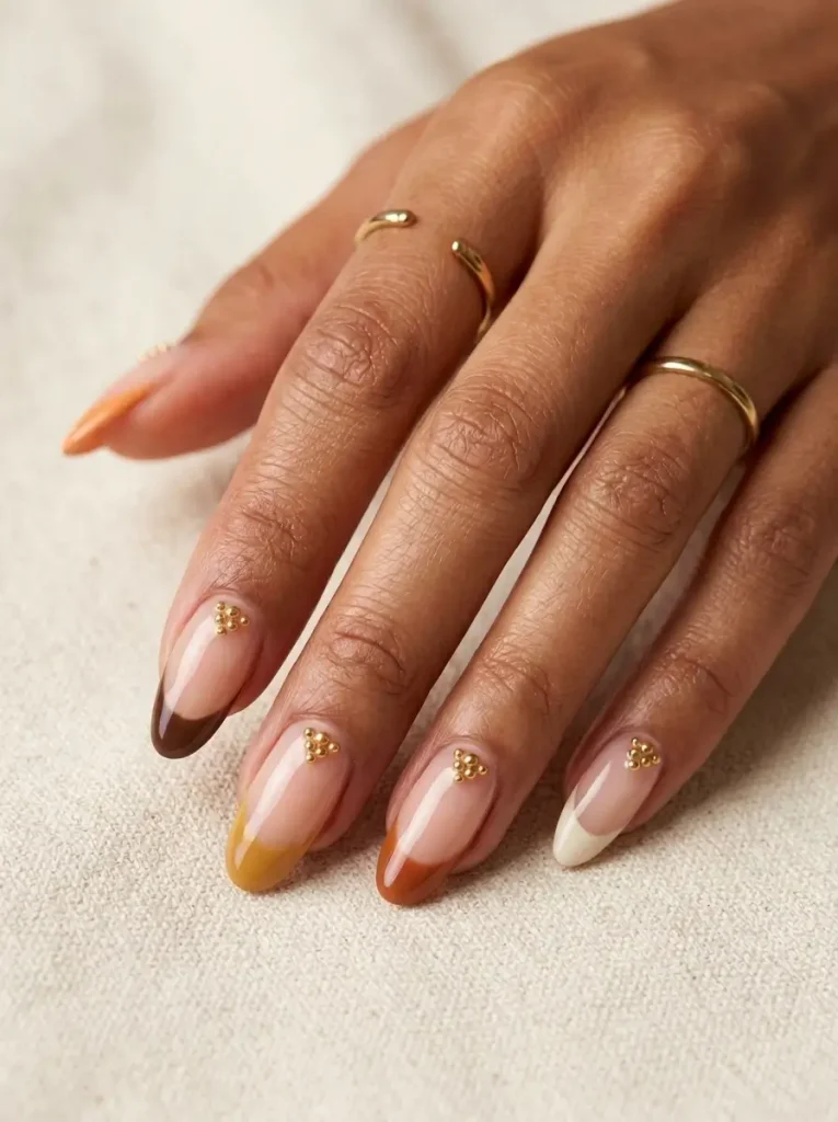 10 Thanksgiving Nail Designs for 2026, From Turkey to Pumpkin Pie 5 Long almond Thanksgiving French tip nails, a different fall tip on each nail with tiny gold beads, on a tan skin tone