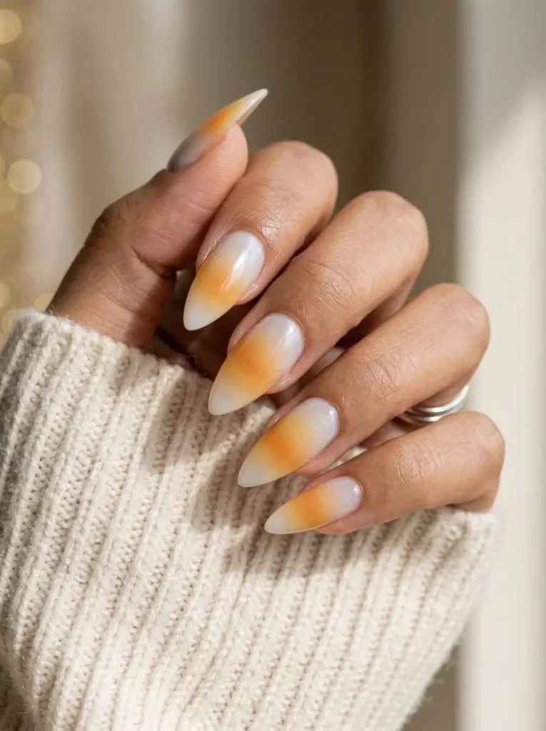 Stiletto nails in a milky pearl glaze with a yellow and orange fade on the tip half and a white point, on an olive skin tone