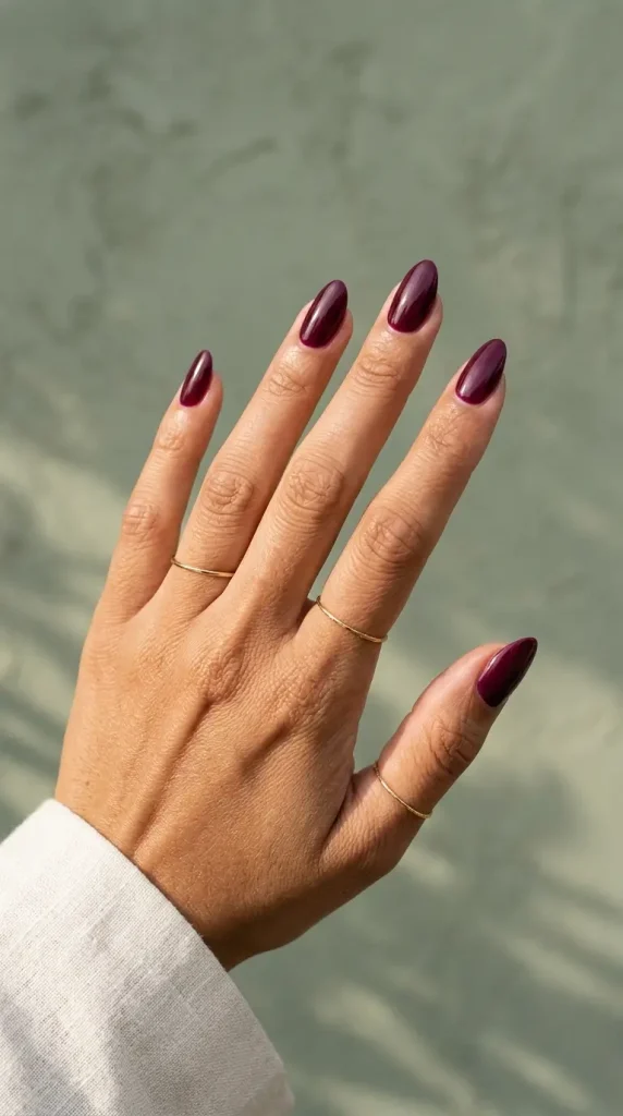 Medium almond nails in glossy deep burgundy polish with a purple undertone, on a tan skin tone