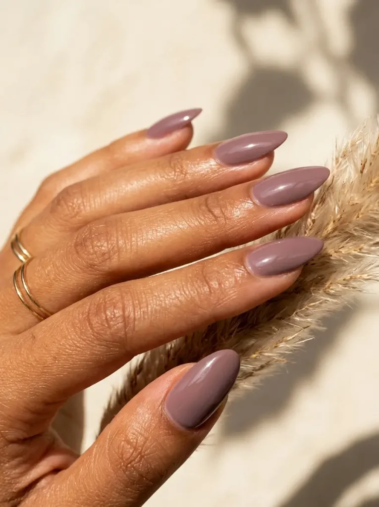 Medium almond nails in glossy dusty mauve polish, a muted pink-purple, on a tan skin tone
