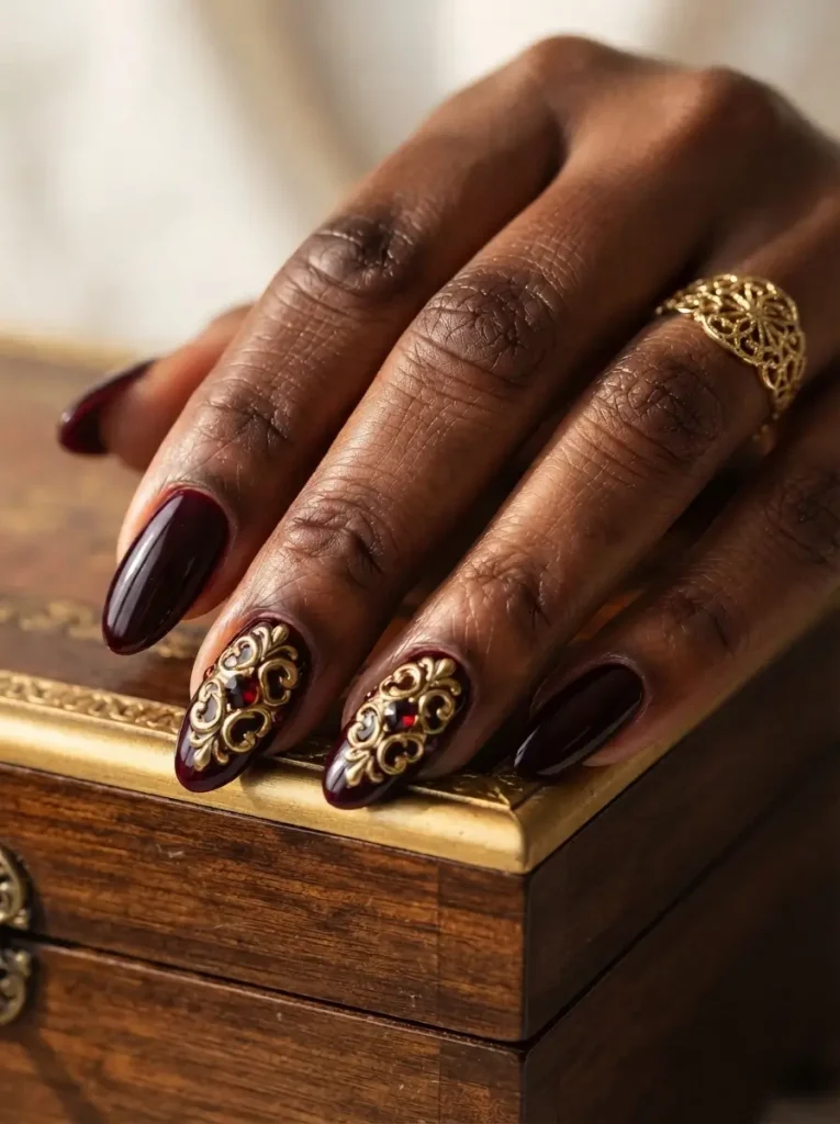Long almond vampire nails in black-cherry red with raised gold baroque scrolls and a red gem on two nails, on a deep skin tone