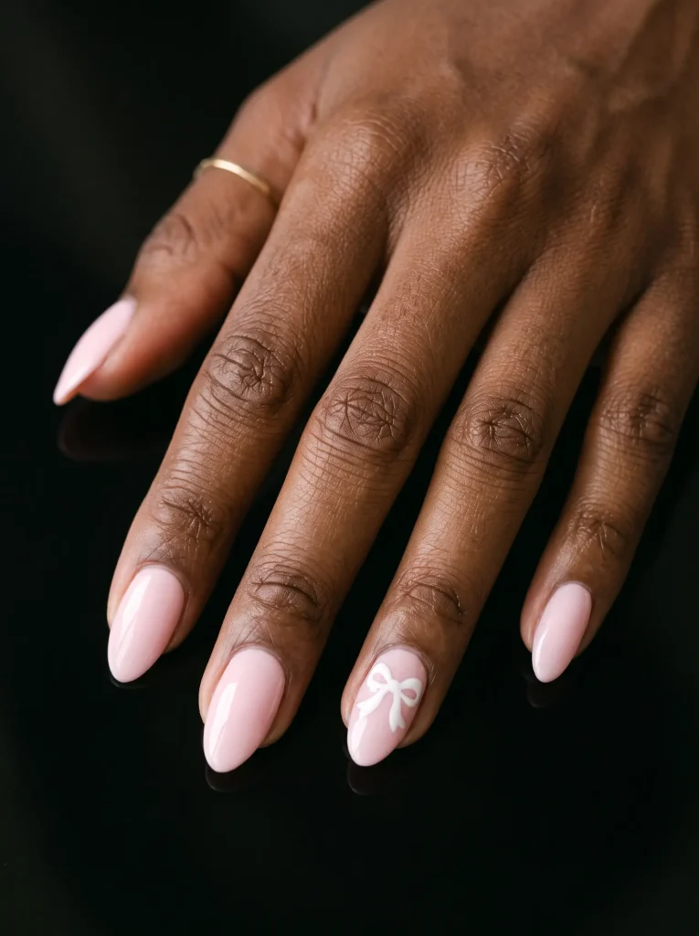 Medium almond pale pink nails with one small white bow painted on a single accent nail and the rest plain, on deep skin