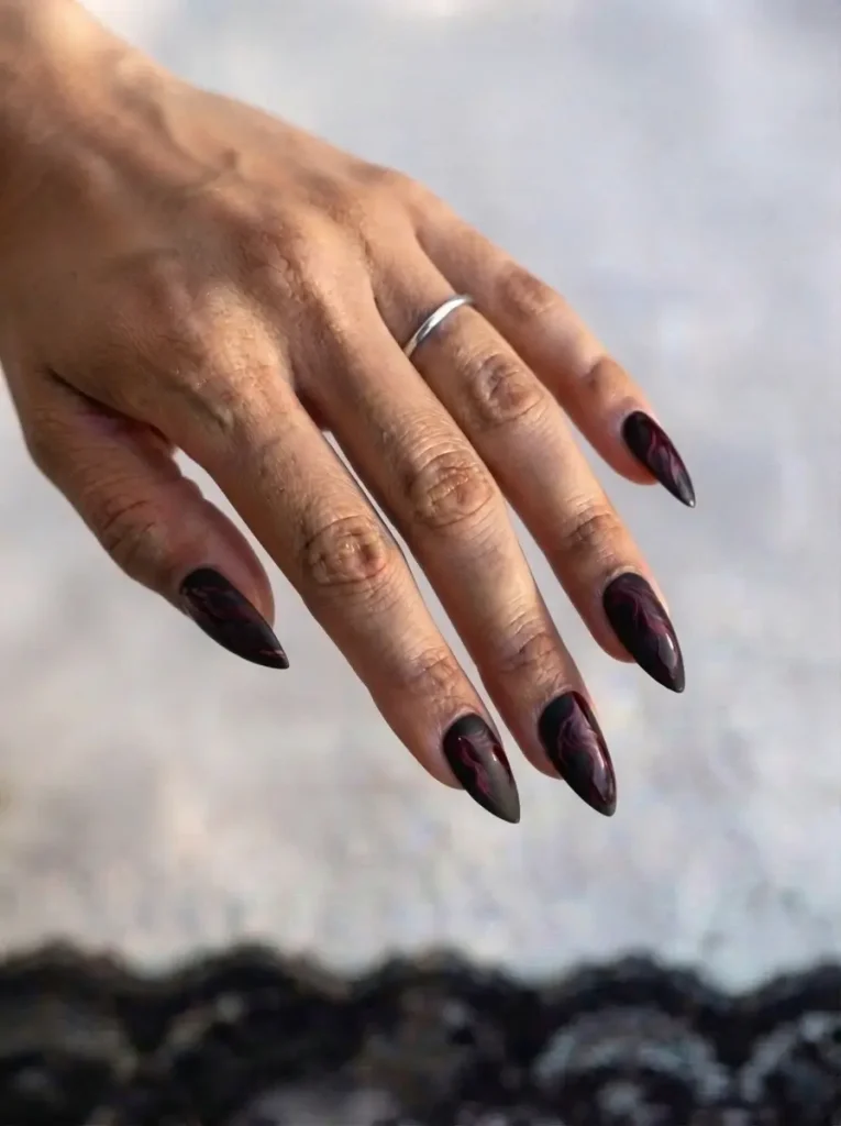 Long stiletto vampire nails in matte black with thin glossy deep red smoke marble veins, on an olive skin tone