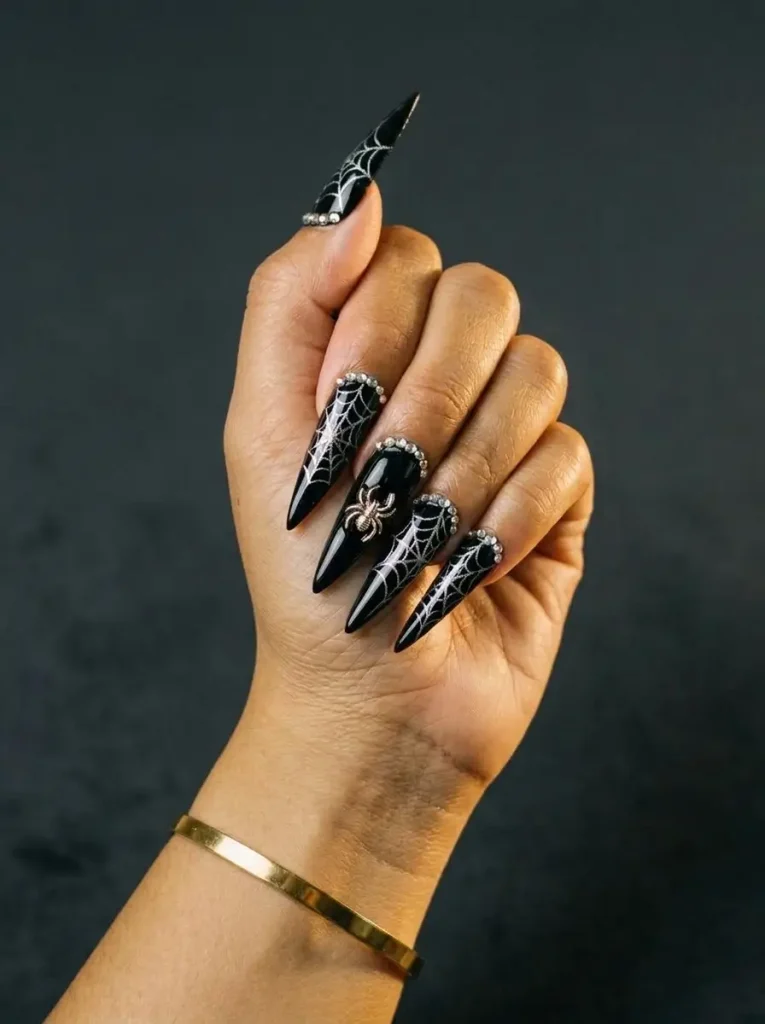 Long black stiletto nails with silver spiderwebs and rhinestones along the cuticle line, on a medium skin tone
