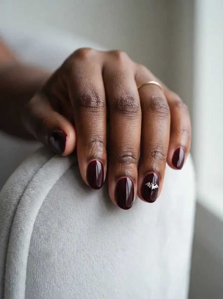 Short oval vampire nails in glossy black cherry with one tiny silver bat charm on the ring finger, on a deep skin tone