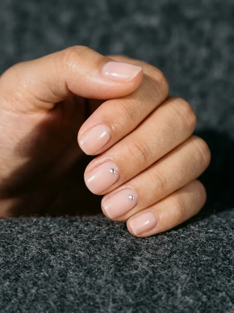 Short round nude nails with one small crystal on two nails and the rest left plain, glossy finish, on medium skin