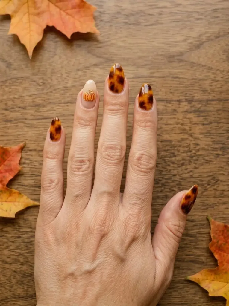 Almond nails in amber tortoiseshell with a small orange pumpkin outlined in gold on one nail, on a light skin tone