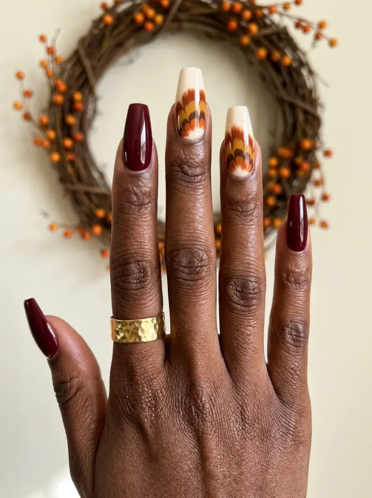 10 Thanksgiving Nail Designs for 2026, From Turkey to Pumpkin Pie 3 Long coffin Thanksgiving nails in burgundy with rust, mustard and brown turkey feathers on two cream nails, on a deep skin tone